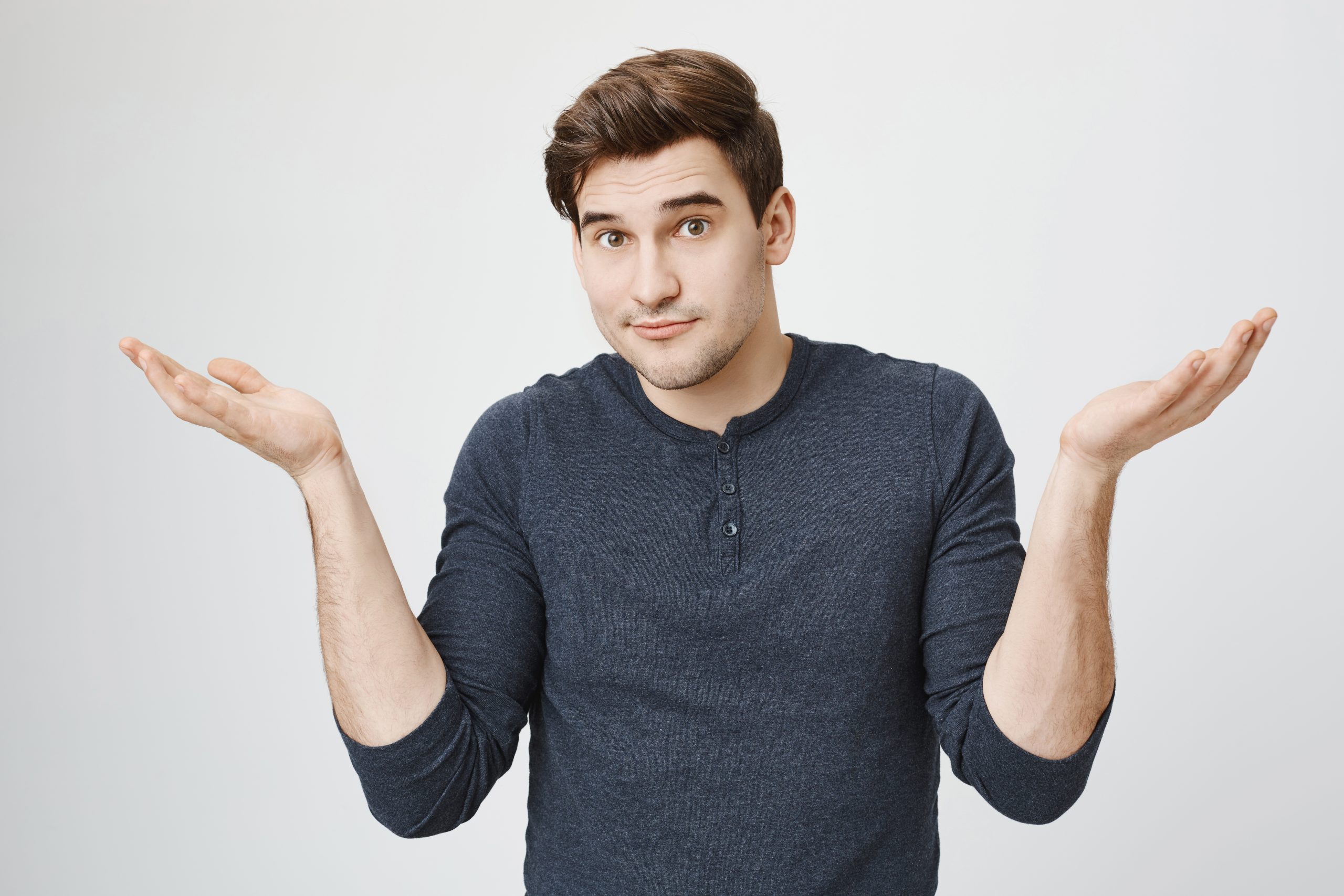Indoor portrait of confused handsome guy showing I have no idea gesture, shrugging shoulders and raising hands, standing against gray background. Sorry I did not know it was your food in fridge