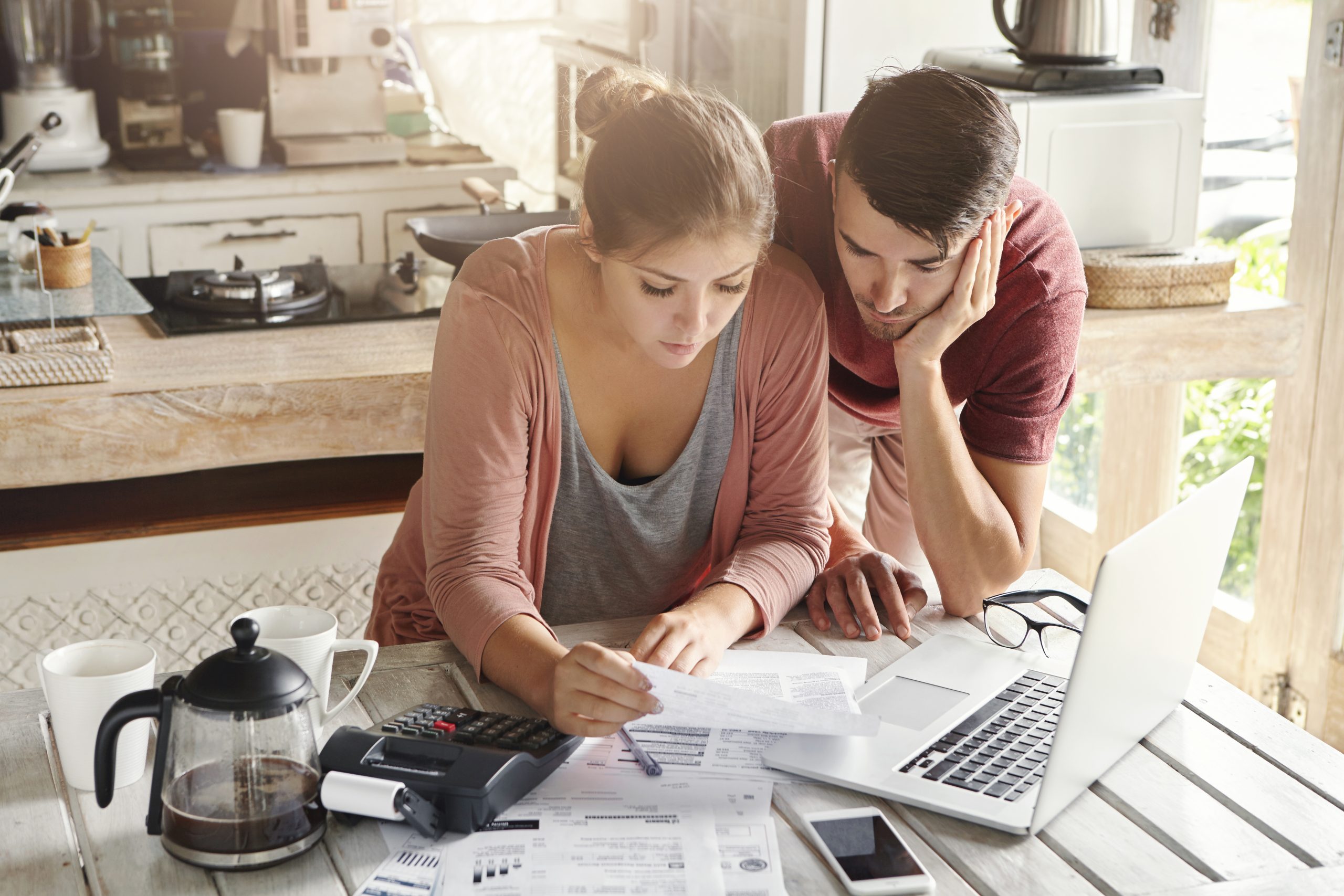 Young stressed family paying utility bills online using laptop. Worried woman holding document, calculating domestic expenses together with her husband, sitting at table with papers and calculator